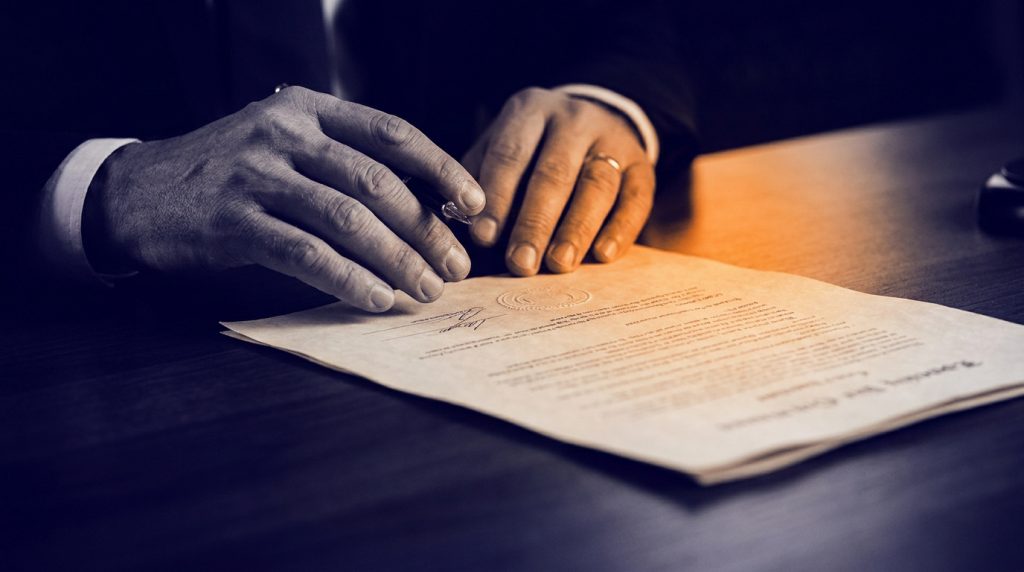 Mains d'une personne en costume tenant un stylo au-dessus d'un document officiel avec signature et sceau, sur un bureau sombre.